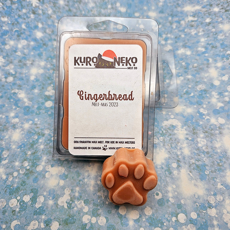 A set of wax melts in a clamshell on a blue speckled background with a paw shaped wax melt in front of it. The pictured scent is Gingerbread.