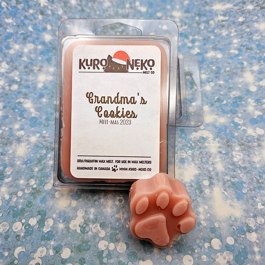 A set of wax melts in a clamshell on a blue speckled background with a paw shaped wax melt in front of it. The pictured scent is Grandma's Cookies.