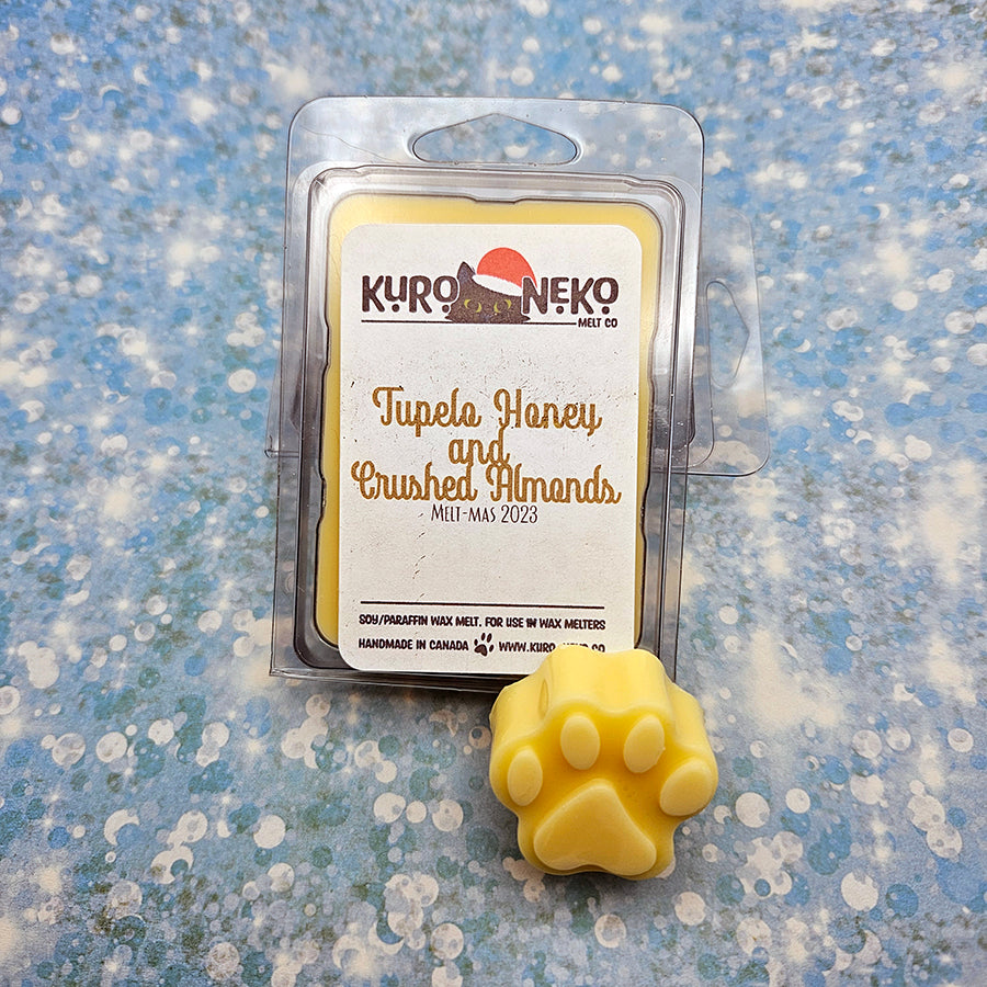 A set of wax melts in a clamshell on a blue speckled background with a paw shaped wax melt in front of it. The pictured scent is Tupelo Honey and Crushed Almonds.